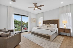 Un dormitorio moderno con una cama grande, un ventilador de techo, una mesita de noche con lámparas, un sofá y vistas al exterior con vegetación.