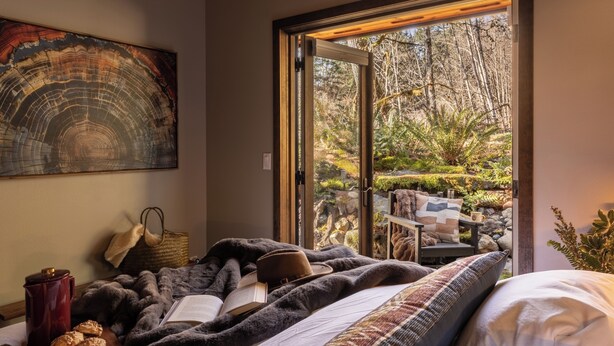 Ein gemütliches Schlafzimmer mit großem Fenster, einem Bett, einer Leseecke mit Buch und Hut sowie einem Bild an der Wand.