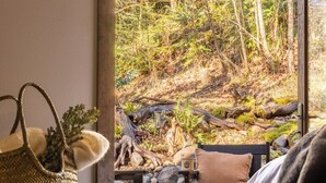 Ein Zimmer mit Holzboden, einem Bett, einem Fenster mit Blick auf einen Wald, einem Tisch mit einer Kamera, einem gewebten Korb und einem Paar Stiefel.