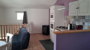 Et kompakt køkken med komfur, ovn og mikroovn. Der er et spiseområde med et bord og stole, og et lille vindue med gardin.