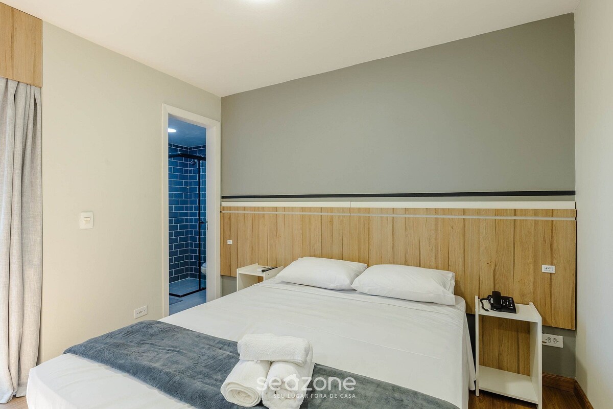A hotel room with a bed, a bedside table, a nightstand, and a bathroom visible through an open door.