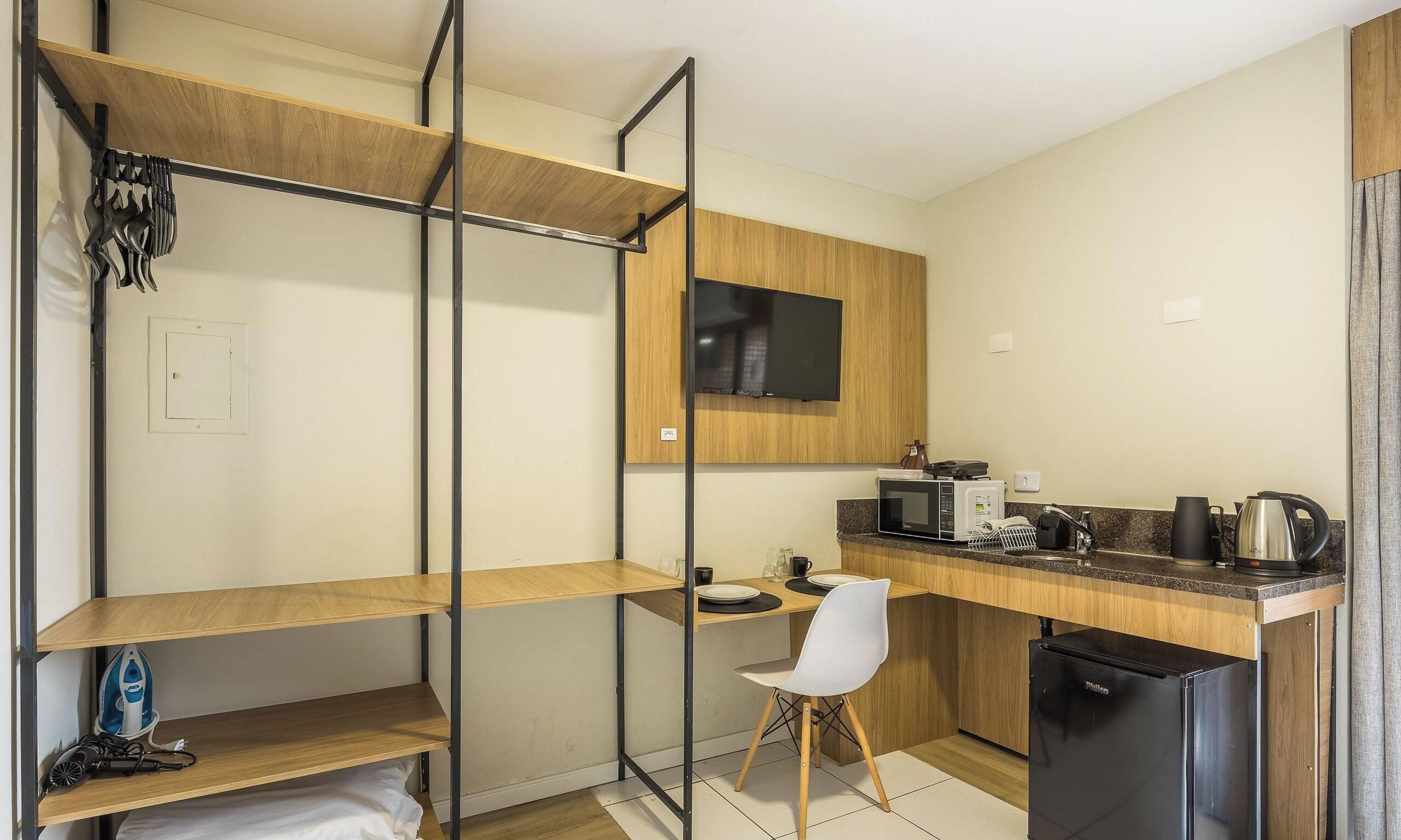 A compact kitchen with a microwave, kettle, and coffee maker on the counter, a small refrigerator, and a single chair.
