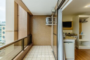 A balcony with a glass railing, a tiled floor, a small kitchen area with a refrigerator, and a television. - Studios at Upper Residence - Upps II (Curitiba)