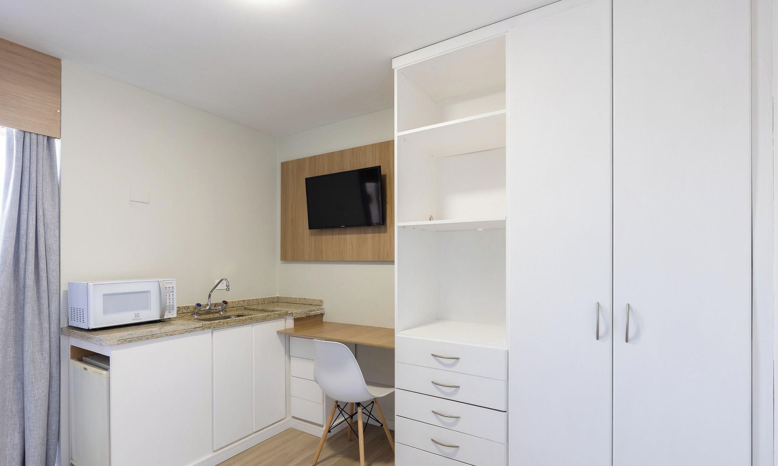 Habitación compacta con kitchenette, televisor de pantalla plana en la pared y un armario blanco con cajones.