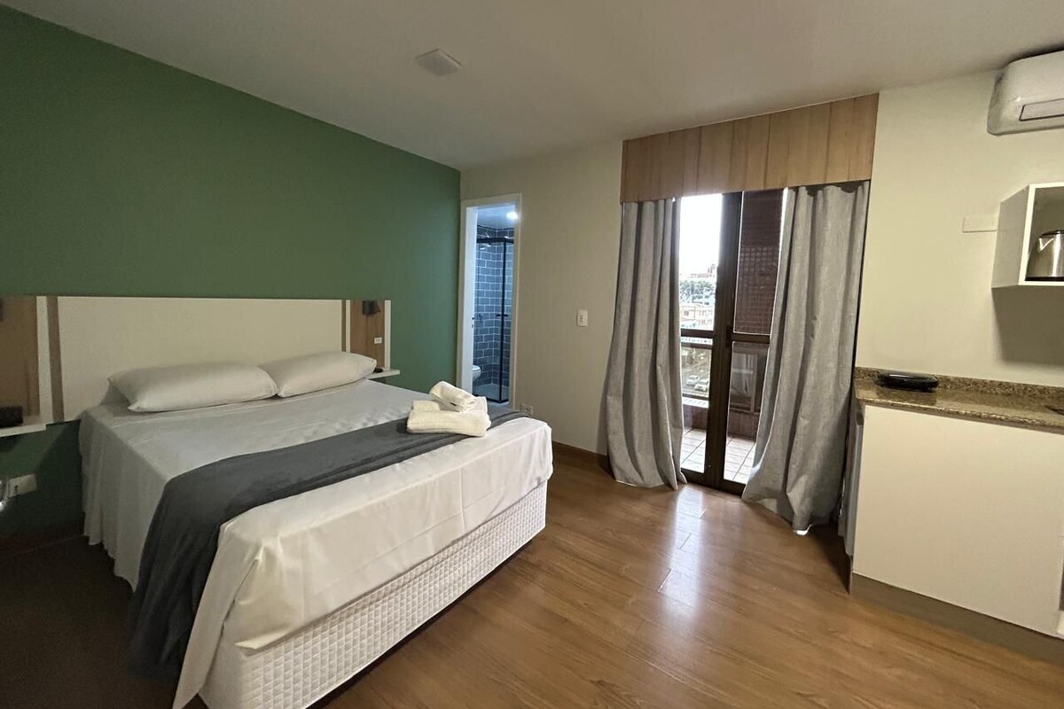 A hotel room with a bed, a small kitchenette, a balcony with curtains, and a view of the outside.