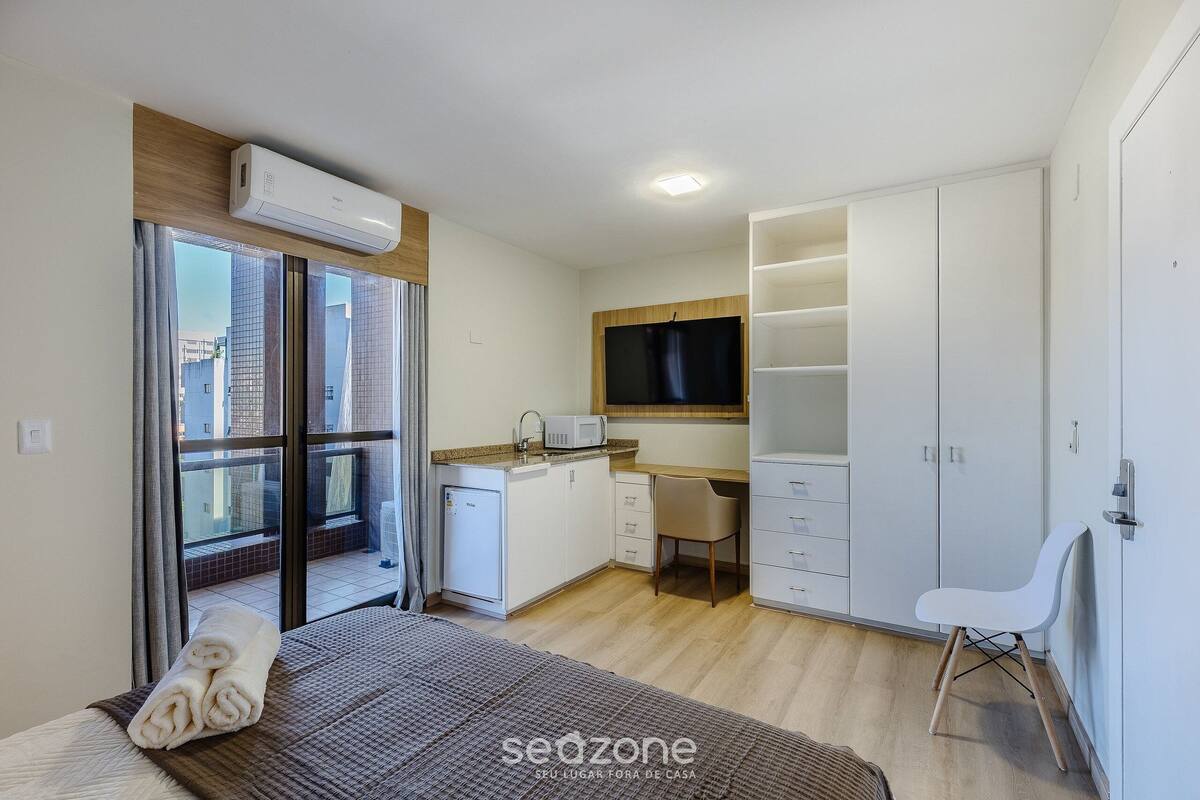 Een slaapkamer met een bed, nachtkastje, raam, schuifdeur naar een balkon en een kitchenette.