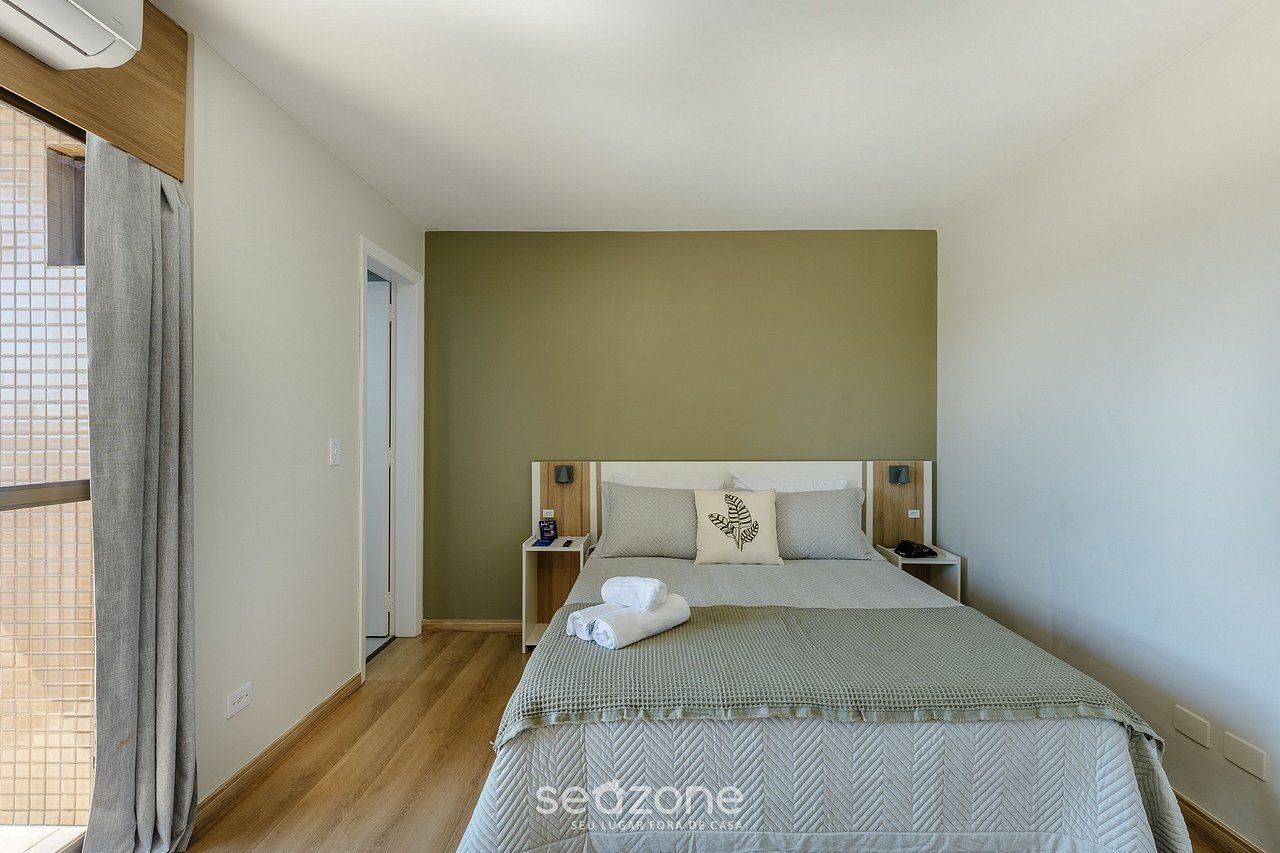 Um quarto com uma cama, duas mesas de cabeceira, um criado-mudo, uma janela com cortinas e uma porta.