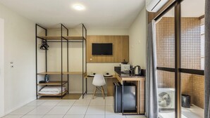 A compact, modern room with a kitchenette, a flat-screen TV mounted on a wooden panel, a desk with a chair, and a sliding glass door leading to a balcony. - Studios at Upper Residence - Upps II (Curitiba)