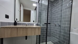 A modern bathroom with a wooden vanity, a glass shower enclosure, and grey tiled walls. - Studios at Upper Residence - Upps II (Curitiba)