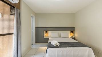 Quarto de hotel com cama, mesa de cabeceira, luminária na parede e janela com cortinas.