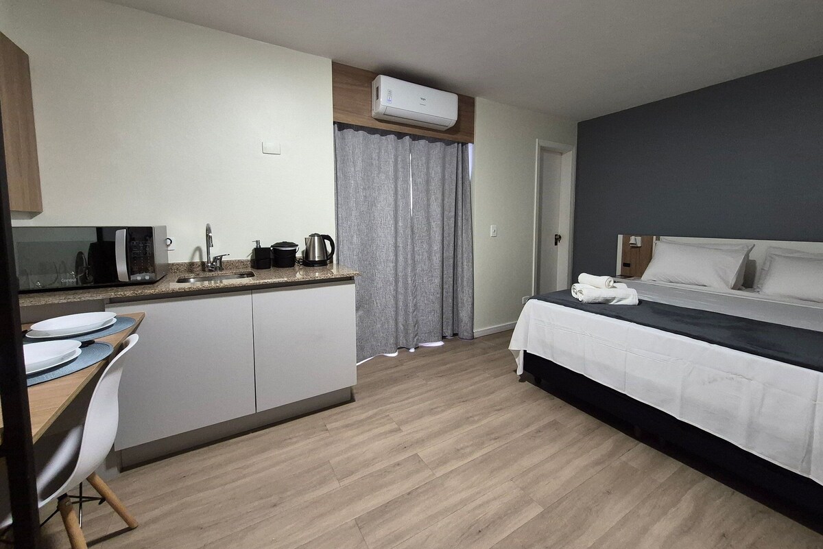 A compact hotel room with a kitchenette, a bed with a dark headboard, and a wooden floor.