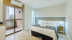 Habitación de hotel con cama, mesita de noche, ventana con cortinas, equipo de aire acondicionado y vista a edificios.