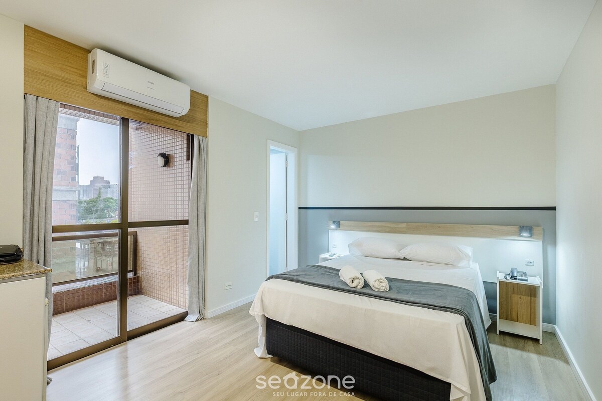 A hotel room with a bed, a nightstand, a window with curtains, an air conditioning unit, and a view of buildings outside.