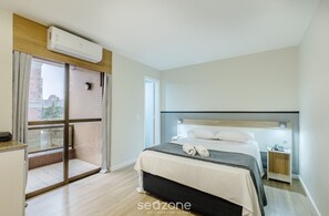 A hotel room with a bed, a nightstand, a window with curtains, an air conditioning unit, and a view of buildings outside.