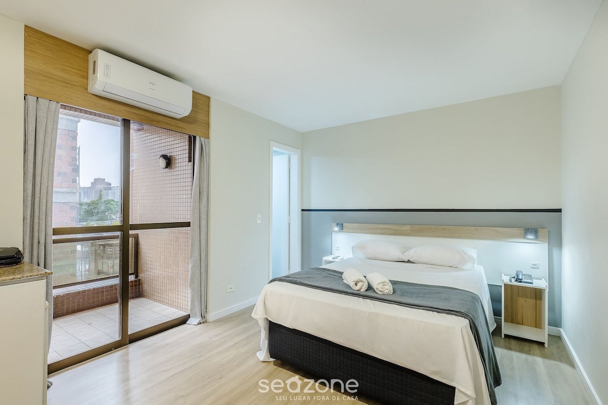 Een hotelkamer met een bed, een nachtkastje, een raam met gordijnen, een airconditioning en uitzicht op gebouwen.
