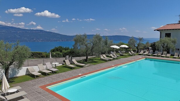 Ein Swimmingpool mit Liegestühlen und Sonnenschirmen, Blick auf einen See, und ein Gebäude mit rotem Dach.