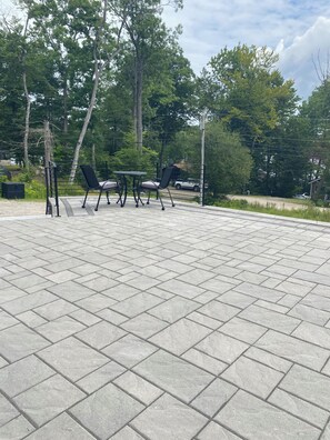 A paved patio area with a table and chairs, surrounded by trees. - Eagle Heights: luxurious Sebago Lake retreat sleeps 8+ (Sebago)