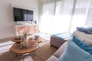 A modern living room with a flat-screen TV, a wooden coffee table with decorative items, a sofa, and a large window with sheer curtains. - Bluez Suites | Pool | Beach | Parking | BBQ (Punta Cana)