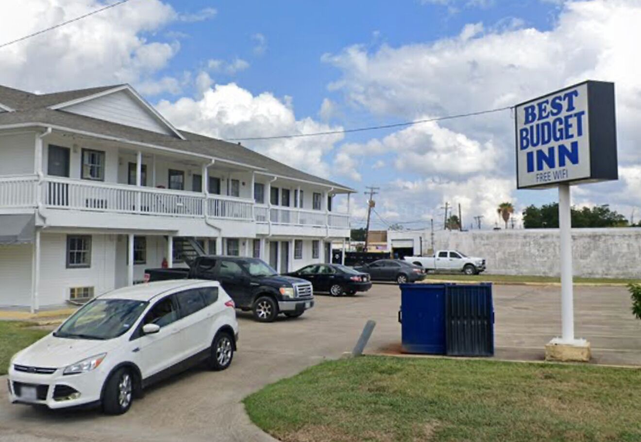 Un motel avec une enseigne indiquant « MEILLEUR RAPPORT QUALITÉ-PRIX » et « WI-FI GRATUIT ». Plusieurs voitures sont garées devant, et une benne bleue est visible.
