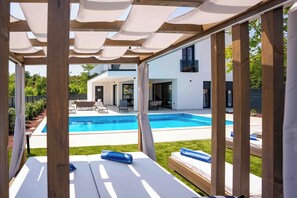 A modern outdoor pool area with a pergola, lounge chairs, and a view of a house. - 5 stars Luxury Villa in Lopar on island Rab (LOPAR (RAB))