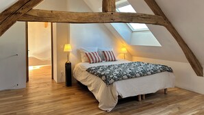 Ett sovrum med ett sluttande tak, träbjälkar, en säng med ett blommigt överkast, två sänglampor och ett takfönster.