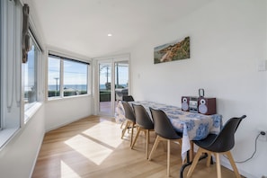 Comedor con mesa preparada para cuatro personas, una radio y vista al mar a través de amplias ventanas.