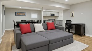 A modern living room with a grey sectional sofa, a dining table with chairs, a kitchen area, and a desk with a chair. - Quiet Area - Revamped Basement (Chicago)