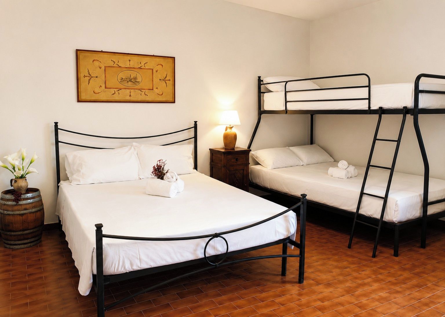 Una habitación con cama individual, literas, un barril de madera y un cuadro enmarcado en la pared.