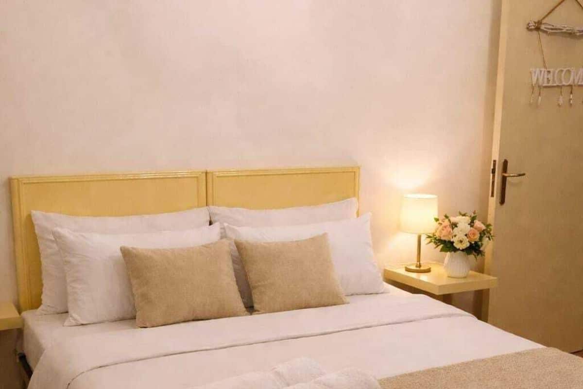 A neatly made bed with white and beige bedding, a bedside lamp, and a welcome sign on the door.