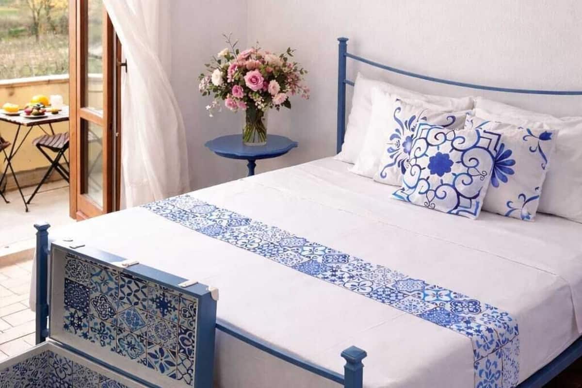 A bedroom with a bed, a nightstand, a vase of flowers, and a view of the outdoors.