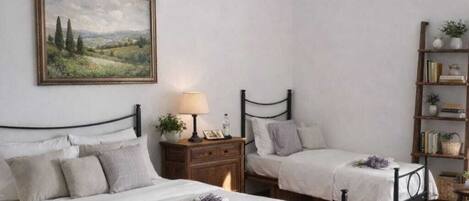 Une chambre avec deux lits, une table de chevet avec une lampe, une bibliothèque et un tableau représentant un paysage, encadré, accroché au mur.