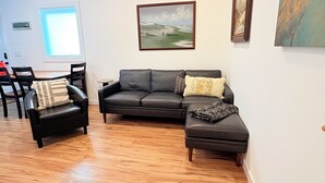 Uma sala de estar com piso de madeira, um sofá de couro preto, uma poltrona de couro preto e um pequeno banquinho. Há um quadro emoldurado na parede e uma janela com persianas.
