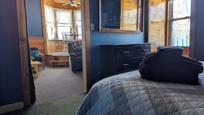 Ein Schlafzimmer mit Bett, Kommode, Stuhl und kleinem Tisch. Ein Fenster mit Vorhängen bietet einen Blick nach draußen.