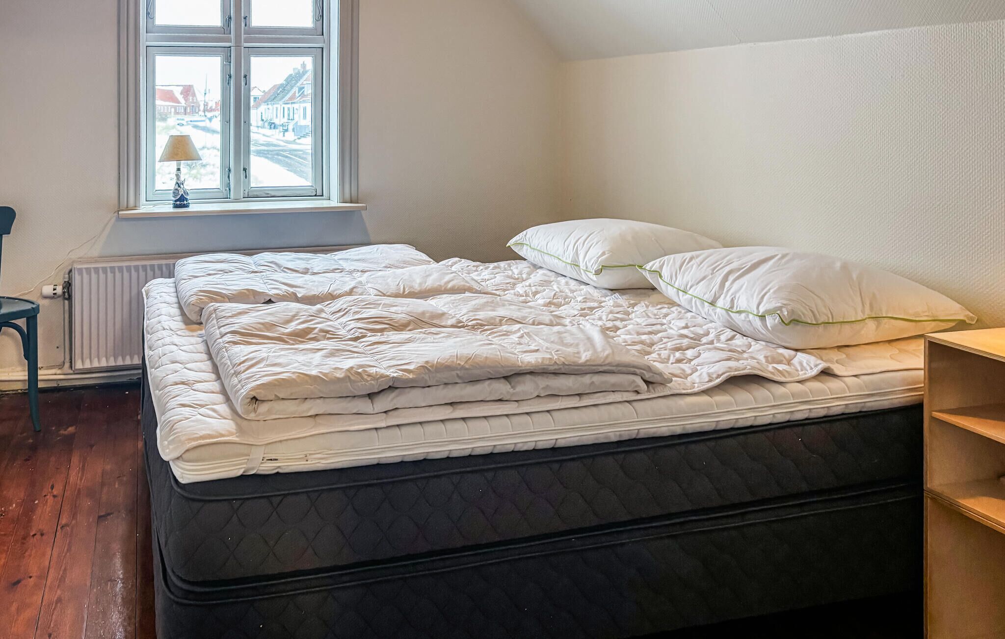Una cama bien hecha con un colchón acolchado y almohadas, una ventana con vistas a edificios, y un radiador en la pared.