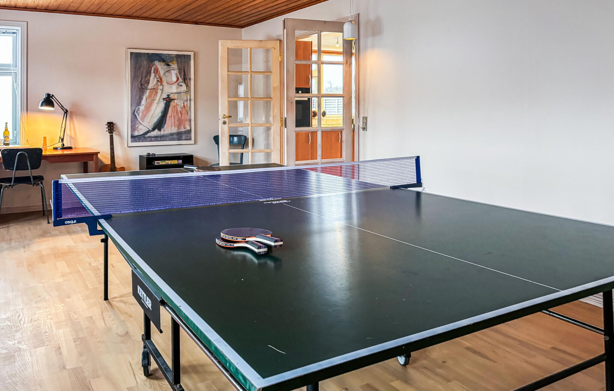 Une table de ping-pong se trouve dans une pièce dont le sol est recouvert de parquet, un tableau est accroché au mur et une porte donne accès à une autre pièce.