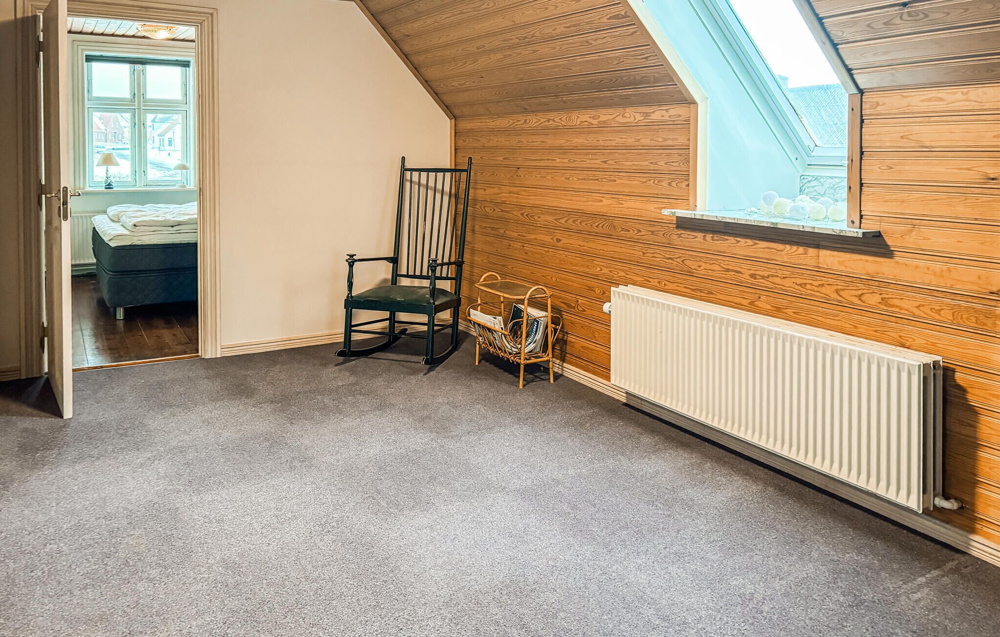 A room with a sloped ceiling, a wooden wall, a radiator, a chair, a magazine rack, and a bed with a view through a window.