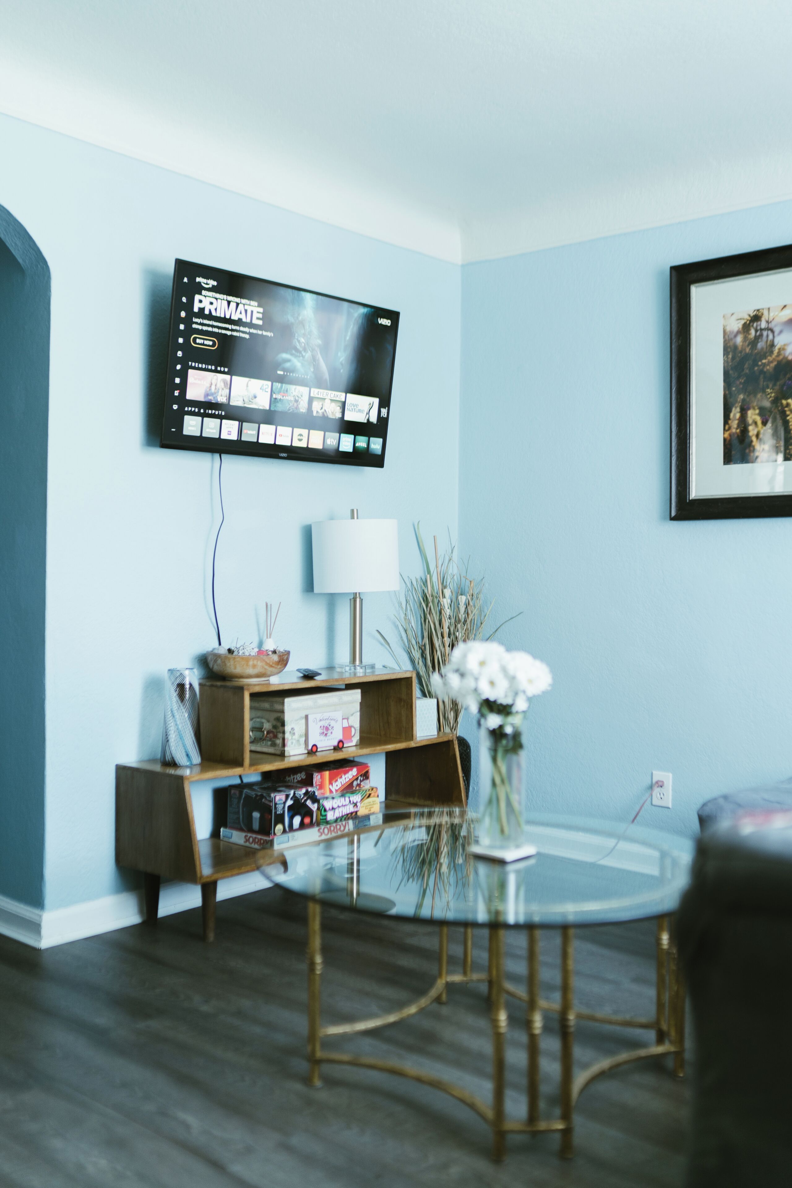 A living room with a mounted TV displaying a movie, a round glass table, a lamp, and a framed picture on the wall.
