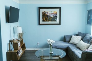 Ein Wohnzimmer mit einem runden Glastisch, einem grauen Sofa, einem gerahmten Gemälde an der Wand und einem an der Wand befestigten Fernseher.