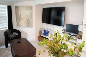 A modern living room with a flat-screen TV, a coffee table, a bookshelf with books and decorative items, and a large abstract painting on the wall. - Stylish Work-Friendly 2BR | Shaw Metro & Dining (Washington)