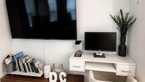 A modern living room with a flat-screen TV mounted on the wall, a smaller TV on a white desk, a bookshelf with books and decorative items, and a comfortable armchair. - Stylish Work-Friendly 2BR | Shaw Metro & Dining (Washington)