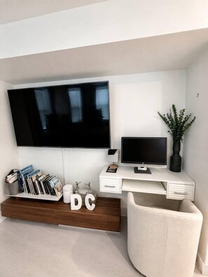 A modern living room with a flat-screen TV mounted on the wall, a smaller TV on a white desk, a bookshelf with books and decorative items, and a comfortable armchair. - Stylish Work-Friendly 2BR | Shaw Metro & Dining (Washington)