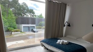 Une chambre avec une grande fenêtre, un lit recouvert d’une couverture bleue, et une vue sur un immeuble et des arbres à l’extérieur.