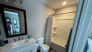Un baño con lavamanos, inodoro y bañera blancos. Hay un espejo sobre el lavamanos y una cortina de ducha sobre la bañera.