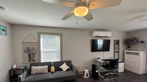 Una sala de estar moderna con un sofá gris, un ventilador de techo, un televisor de pantalla plana, un escritorio con una silla y un equipo de aire acondicionado.