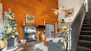 A cozy log cabin interior with a fireplace, a Christmas tree, a dining area, and a staircase. - Adorable 2-bedroom condo cabin with AC in lively Branson close to everything! (Branson)