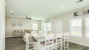 Amplia sala de estar con un sofá blanco grande, una zona de comedor con una mesa de mármol y sillas blancas, y un área de cocina con un cartel que dice 'Cocina' sobre las ventanas.