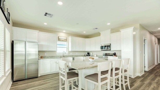 Una cocina moderna con armarios blancos, un refrigerador de acero inoxidable, encimera de granito y un comedor con sillas blancas.