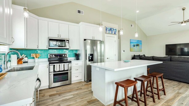 A modern kitchen with white cabinets, a large island with stools, stainless steel appliances, and a teal backsplash. - Teal Turtle - Sassy & Sophisticated Sea Isle Spot (Galveston)
