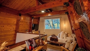 Een gezellig chaletinterieur met houten wanden, een bed met wit beddengoed, een kleintje tafeltje met een plant en een raam met uitzicht op bomen.