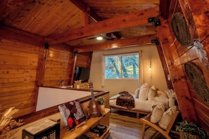 A cozy cabin interior with wooden walls, a bed with white bedding, a small table with a plant, and a window with a view of trees. - Arrowbear Lake: Luxe Cabin: Large Deck & Fire Pit, Workspace & Loft (Arrowbear Lake)